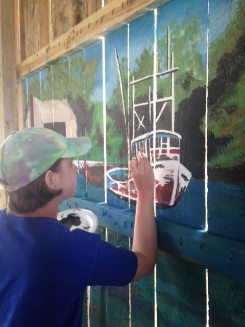 Young artist painting a boat mural on wooden fence